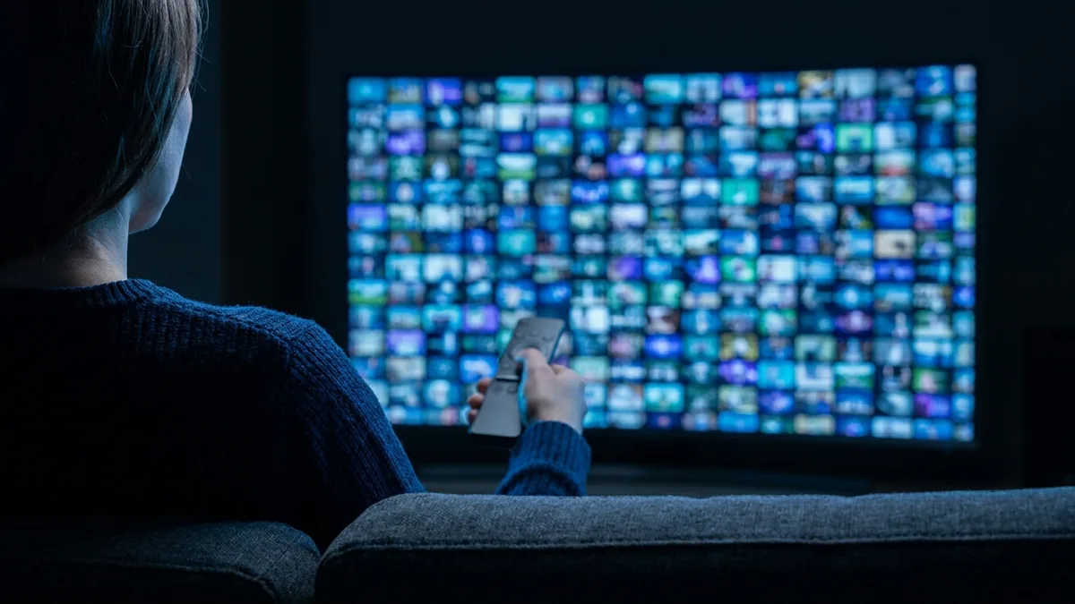 A person holding a remote toward a large television filled with a colourful mosaic of video thumbnails in a dark room